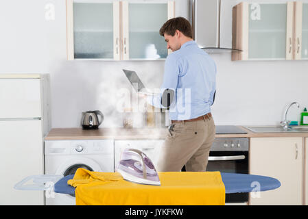 Ferro da stiro a vapore su stoffa mentre l'uomo utilizzando laptop in cucina Foto Stock