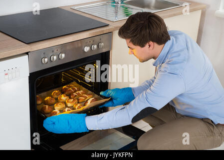 Giovane uomo prendendo vassoio da forno con pane appena sfornato dal forno in cucina Foto Stock