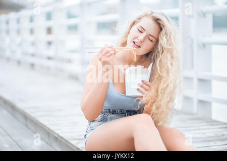 Foto di donna con lunghi capelli ricci, seduta sul pavimento in legno, tenendo una scatola di pranzo e mangiare tagliatelle cinesi take-out con bacchette. Foto Stock