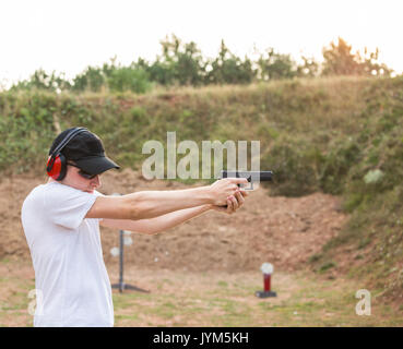 Bel giovane agente di poliziotto di mira e spara contro un bersaglio con una pistola glock desert eagle pistola indossando cappello nero e la camicia bianca in natura ambien Foto Stock