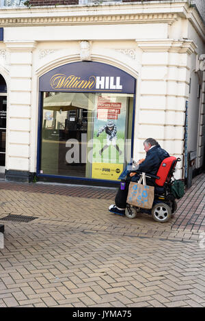 Uomo anziano con un abbigliamento informale che viaggiano su un grigio pavimentazione a spina di pesce passato di equitazione a William Hill Scommesse shop Foto Stock
