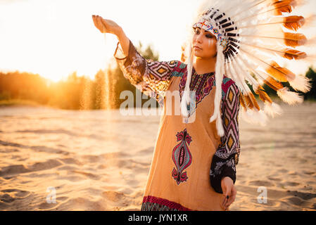 Bella donna in un abito di indiani americani gioca con la sabbia sul tramonto. Giovane ragazza nel copricapo fatto di piume di uccelli selvatici Foto Stock