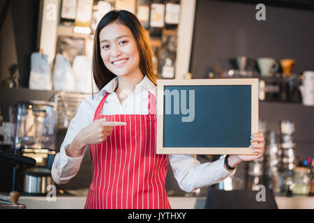 Femmina asiatica barista usura grembiule rosso punto alla lavagna vuota nel menu di caffè al banco bar con sorriso faccia,servizio di caffetteria concetto,lasciano spazio per aggiunta di y Foto Stock