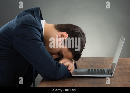 Vista laterale di esaurito imprenditore appoggiato sulla scrivania con computer portatile contro uno sfondo grigio Foto Stock