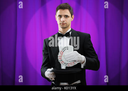 Ritratto di giovane mago mettendo le carte da gioco in hat contro la tenda viola Foto Stock