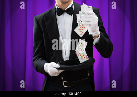 Sezione mediana del mago maschio di eseguire trick con carte e hat contro la tenda viola Foto Stock