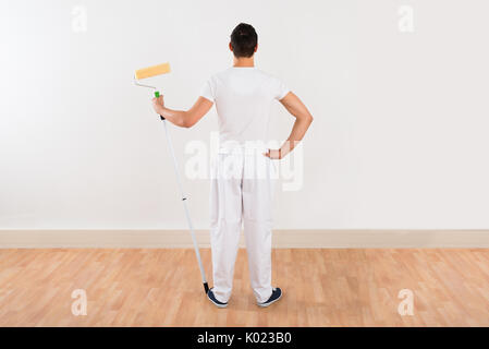 Vista posteriore del giovane azienda rullo di vernice mentre in piedi contro il muro bianco a casa Foto Stock