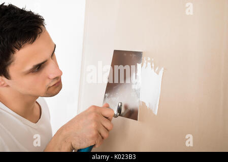 Giovane uomo mediante spatola sul muro bianco a casa Foto Stock