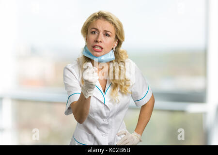 Angry Young nurse puntando con il dito. Femmina frustrato medico o infermiere in ospedale lo sfondo della finestra. Foto Stock