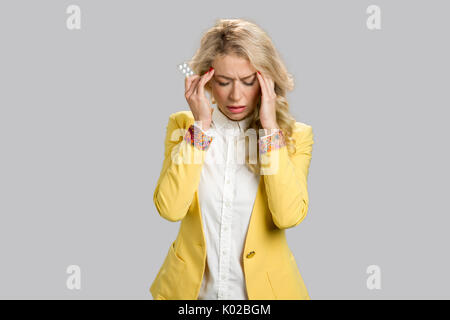 Donna con mal di testa tenendo la medicina. Ritratto di malati giovane donna indossando giacca gialla aventi un forte mal di testa su sfondo grigio. Foto Stock