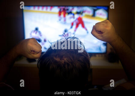 Vista posteriore di emozioni uomo con gesti delle mani in alto come la visione di hockey su ghiaccio in televisione a casa. Vista posteriore della testata maschio a guardare lo sport in TV. Foto Stock