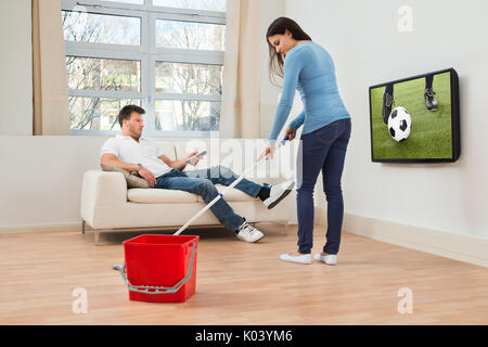 Donna pulizia pavimento davanti all uomo guardando la partita di calcio in televisione Foto Stock