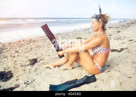 Donna bionda seduto sulla spiaggia con pinne e occhiali da nuoto Foto Stock
