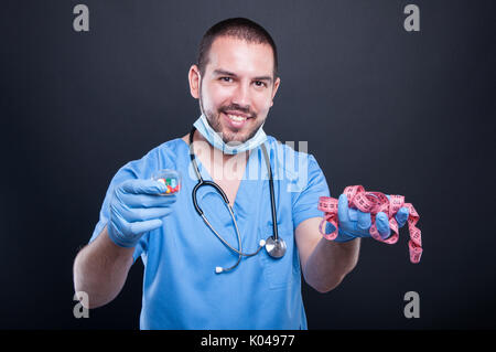 Chirurgo di plastica che indossa scrubs tenendo premuto il nastro di misurazione prima e pillole e sorridente su sfondo nero Foto Stock