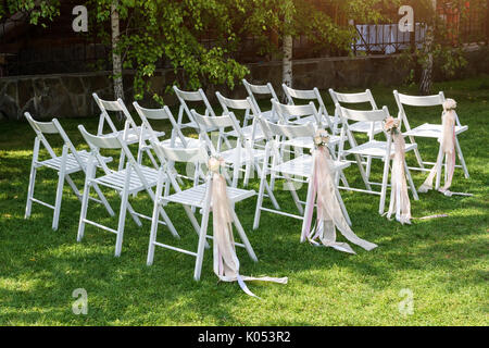 Bellissimo matrimonio istituito. Cerimonia di matrimonio nel giardino. Bianco sedie in legno decorato con fiori e nastri rosa in piedi in righe. Foto Stock