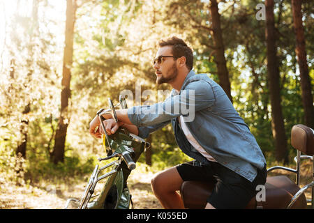 Immagine del bel giovane uomo barbuto seduto su uno scooter all'esterno. Guardando a parte. Foto Stock