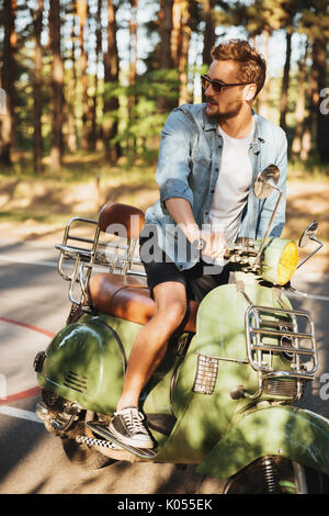 Immagine di concentrato di giovane uomo barbuto seduto su uno scooter all'esterno. Guardando a parte. Foto Stock