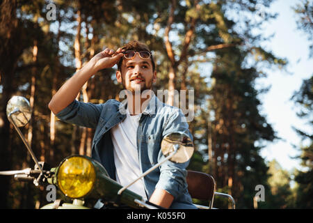 Immagine di grave giovane uomo barbuto seduto su uno scooter all'esterno. Guardando a parte. Foto Stock