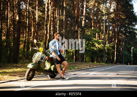 Immagine di grave giovane uomo barbuto in piedi accanto a scooter all'esterno. Guardando a parte. Foto Stock