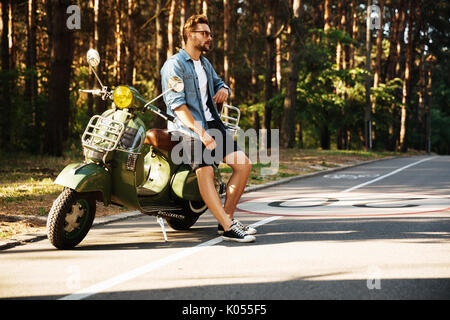 Immagine di grave giovane uomo barbuto in piedi accanto a scooter all'esterno. Guardando a parte. Foto Stock