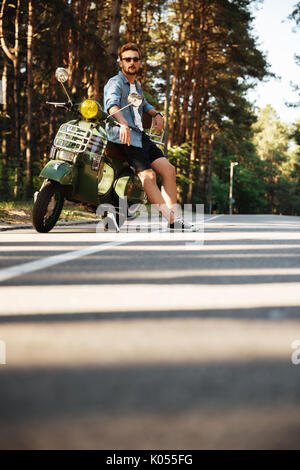 Immagine di concentrato di giovane uomo barbuto in piedi accanto a scooter all'esterno. Guardando a parte. Foto Stock