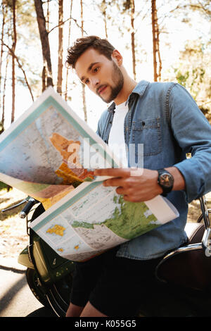 Immagine del bel giovane uomo barbuto vicino a scooter guardando la mappa all'esterno. Foto Stock