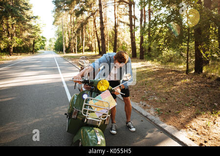 Immagine di grave giovane uomo barbuto in piedi accanto a scooter all'esterno. Guardando a parte. Foto Stock