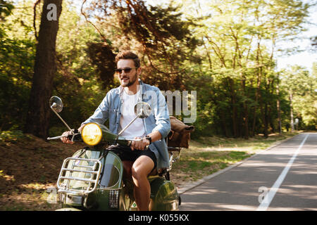 Immagine di grave giovane uomo barbuto su scooter all'esterno. Guardando a parte. Foto Stock