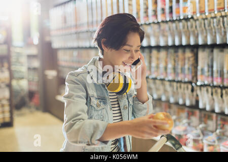 Sorridente giovane donna con cuffie parlando al cellulare a fare la spesa nel mercato Foto Stock