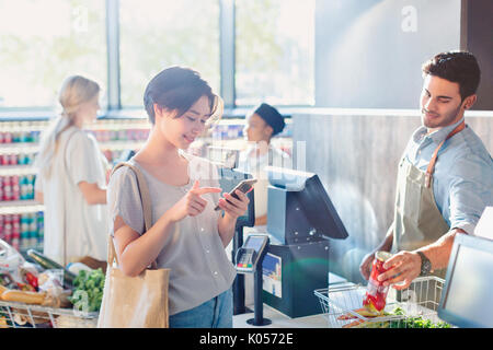 Giovane donna tramite telefono cellulare al negozio di alimentari checkout del mercato Foto Stock