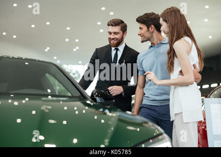 Bel giovane venditore auto raccontare una coppia giovane circa le caratteristiche della vettura presso la concessionaria Foto Stock