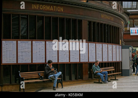 Due uomini ragazzi seduti sui banchi del treno tabelle tempo sfondo utilizzando smart phone sotto in pannelli di legno segno informazioni la stazione centrale di Glasgow concourse Foto Stock