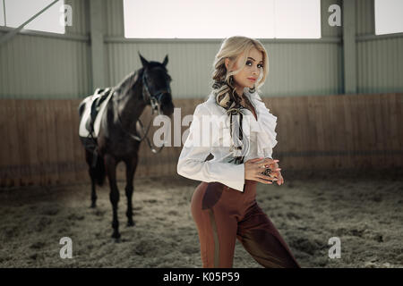 Bellissima ed elegante ragazza bionda sta in piedi vicino al suo cavallo nero medicazione di concorrenza uniforme camicetta bianca maglietta e pantaloni marrone. Piscina portraite in Foto Stock