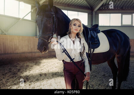 Bellissima ed elegante ragazza bionda sta in piedi vicino al suo cavallo nero medicazione di concorrenza uniforme camicetta bianca maglietta e pantaloni marrone. Piscina portraite in Foto Stock