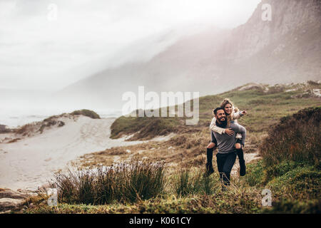 Colpo all'aperto dell'uomo che donna piggyback sulla spiaggia invernale. Uomo Donna portante oh la schiena rivolta lontano. Foto Stock