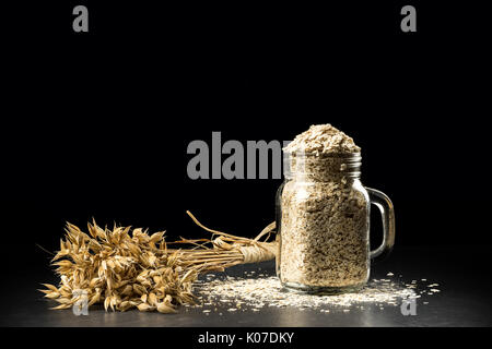 Grappolo d'avena e fiocchi nel vasetto di aromatizzanti, isolato su sfondo nero. profumo di grano, golden avena spikelets scuro sul tavolo di legno, può essere riempito con secchi Foto Stock