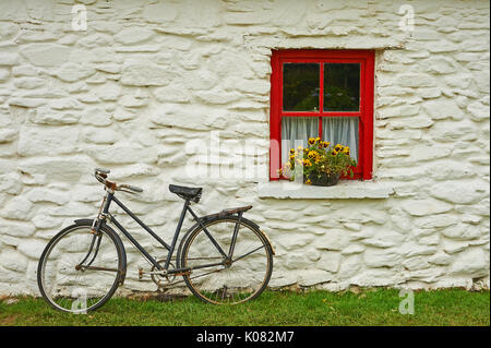 Una vecchia bicicletta appoggiata contro un lime lavato costruzione in pietra con un dipinto di rosso finestra e fiori di colore giallo Foto Stock