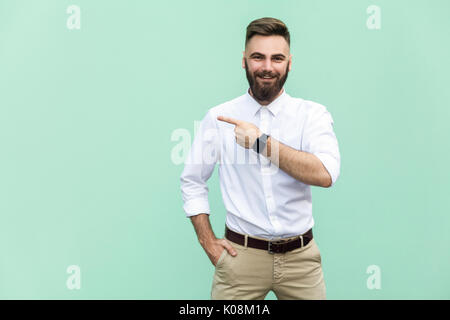 Imprenditore copia di puntamento dello spazio. Bel giovane uomo adulto con la barba in maglia bianca guardando la fotocamera e rivolto lontano rimanendo isolate su lig Foto Stock