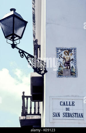 Una illustrazione di religiose di piastrelle di ceramica segna Calle de San Sebastian, una strada chiamata dopo inizio martire cristiano San Sebastiano nella storica Vecchia San Juan di Porto Rico (PR), un territorio non costituite in società degli Stati Uniti nel Mar dei Caraibi. Una delle piastrelle che mostra le spoglie del santo della parte inferiore delle gambe è mancante. Un'annata del ferro battuto via la luce illumina il nome della strada ed illustrato e segno di notte. Foto Stock