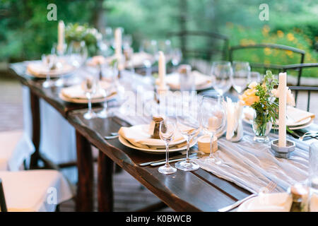 Fest, cena, tradizione concetto. lunga tavola quercino servita per la celebrazione con posate, piatti e trasparente gli occhiali di abbagliamento, fiori e candele in interessanti i titolari in forma di pianta ottagonale Foto Stock