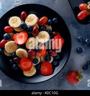 Utile prima colazione di frutti e bacche, vista dall'alto. Fette di banane, fragole, uva e mirtilli in una piastra nera Foto Stock