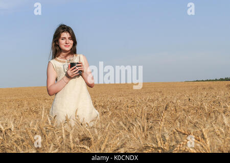 Giovane donna attraente in splendido abito naturale a piedi con con un bicchiere di birra scura nel golden campo di grano durante la luce del sole. Foto Stock