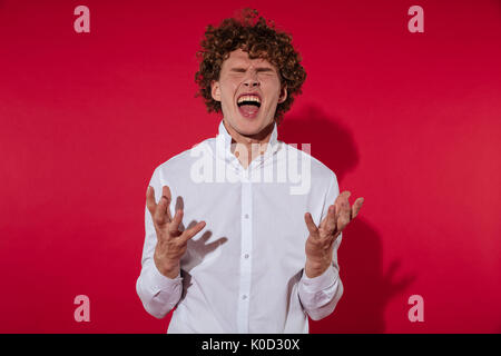Eccitato Giovane Uomo in camicia urlando con gli occhi chiusi e gesticolando isolate su sfondo rosso Foto Stock