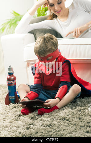 Carino ragazzo supereroe di giocare ai videogiochi seduta sul tappeto con sua madre sullo sfondo. Foto Stock
