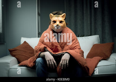 Giovane uomo che indossa una maschera di fox seduto sul divano coperto con una coperta. Foto Stock