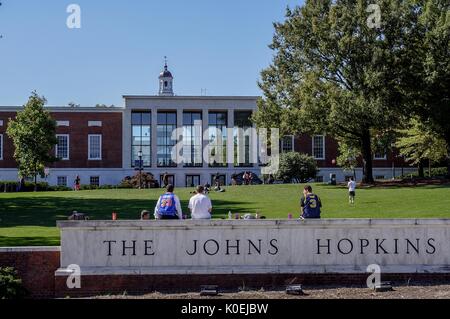 Vista da Charles Street della 'Beach' erbosa e della Biblioteca Milton S Eisenhower della Johns Hopkins University; tre studenti maschi, le loro spalle di fronte alla macchina fotografica, siedono sulla cima del segno di marmo dell'università, altri studenti prendere il sole e giocare a cattura sulla collina erbosa; La torre dell'orologio di Gilman Hall, un edificio di scienze umane, si inarsa sopra la cima della biblioteca; Baltimora, Maryland, marzo 2014. Cortesia Eric Chen. Foto Stock