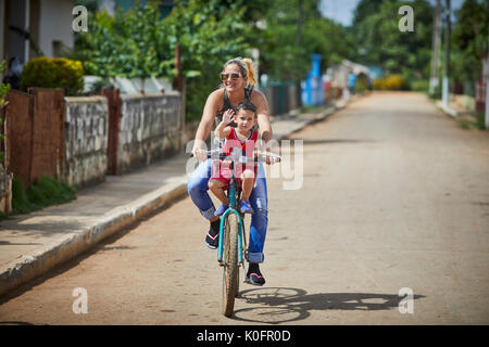 Cubano, Cuba, Cardenas, madre cavalca il suo bambino su una bicicletta attraverso il villaggio Foto Stock