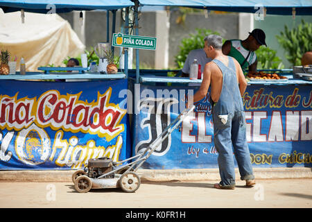 Cubano, Cuba, Cardenas, uomo spingendo un tosaerba attraverso le strade di un villaggio di poveri a sulla pannocchia di mais in stallo Foto Stock