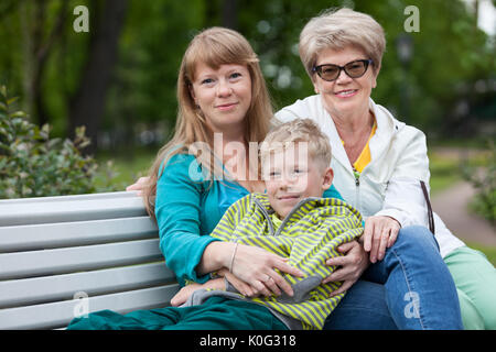 Famiglia di tre persone cercano in telecamera seduto sulla panchina da giardino insieme in abbracci, ragazzo, adulti e anziani le donne Foto Stock