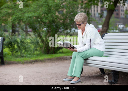 Senior donna legge libro seduti sul bordo della bianca lunga panca di legno in estate park, spazio di copia Foto Stock